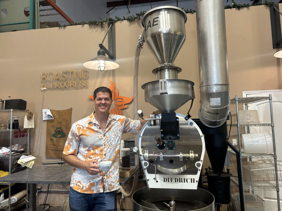 Man stands in a Hawaiian shirt in front of a large coffee roaster