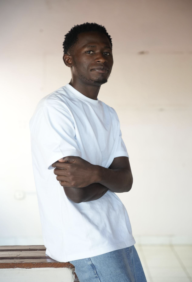 A young man leans against something with arms crossed, looking into the camera