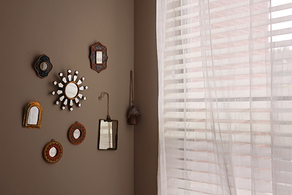 Seven miniature mirrors with interesting designs and borders hang in a cluster on a wall adjacent to a window.