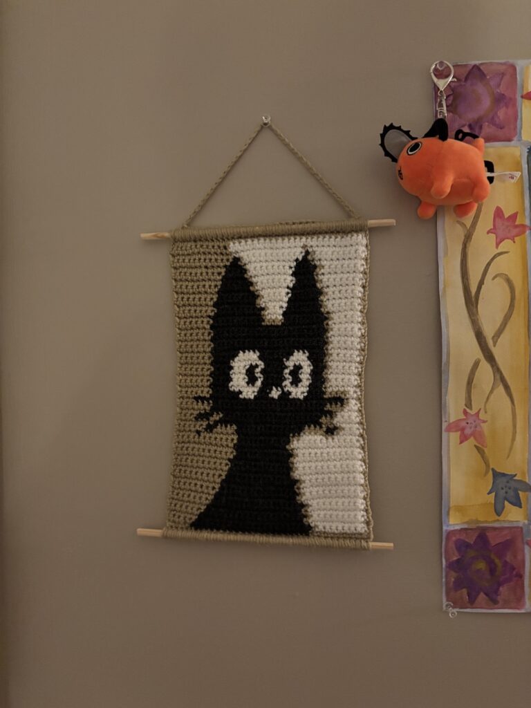 A crochet tapestry of a black cat hangs on a wall.