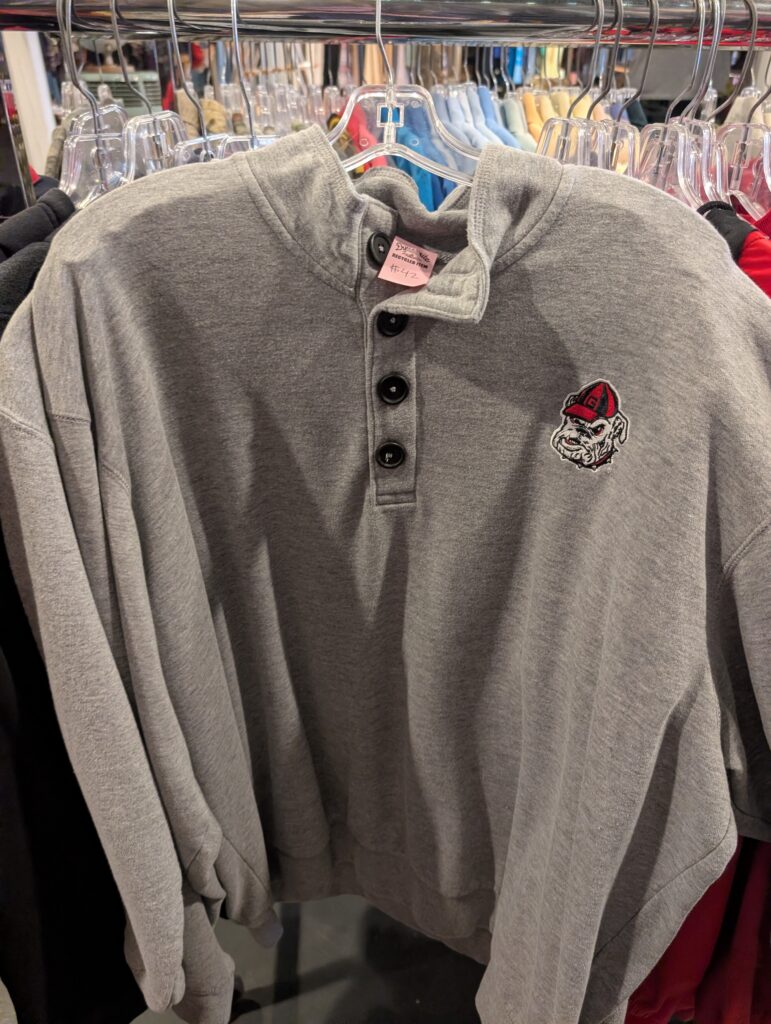 A grey henley sweatshirt with a Georgia Bulldogs logo hangs on a rack inside of Dynamite Clothing, with a tag pricing the sweatshirt at $42. 