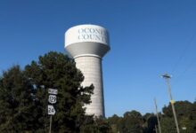 A white water tower with the words "Oconee County" on it behind some trees.