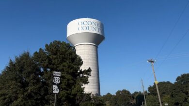A white water tower with the words "Oconee County" on it behind some trees.