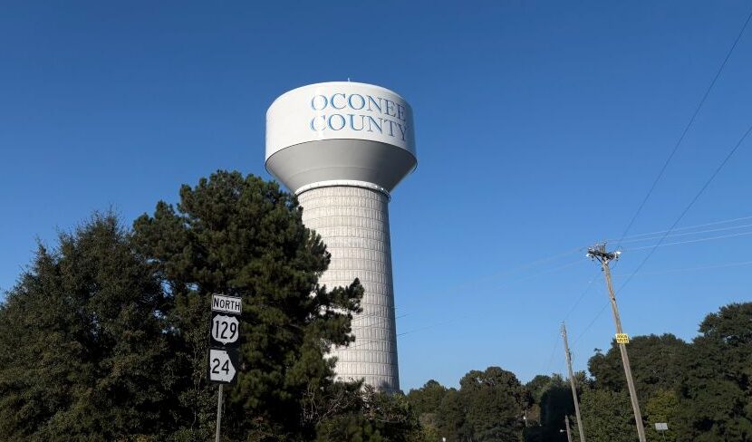 A white water tower with the words "Oconee County" on it behind some trees.