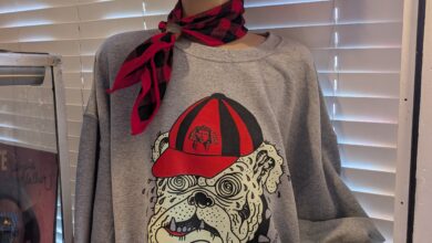 A mannequin head sits at the entrance of Dynamite Clothing, wearing a grey Georgia Bulldogs sweatshirt the store designed and sells, while wearing a scarf and pink earmuffs, resting above a sign that says "No Returns".